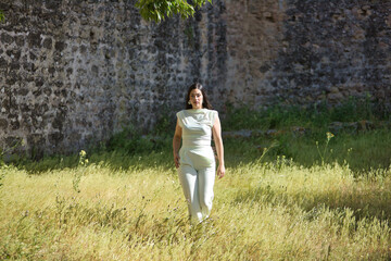 Young, pretty, brunette woman in a white dress, relaxed and calm, walking in the tall grass on a...