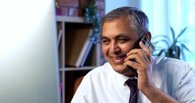 Indian executive working on computer while attending phone call at desk in modern office, Asian corporate professional multitasking with focus and communication skills in contemporary workplace setup