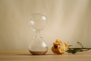 A simple hourglass on a wooden table with dry rose a beige backdrop, symbolizing time flow, aging,...