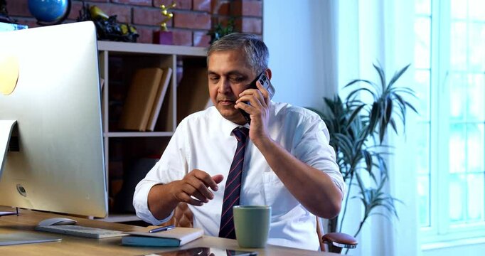 Indian executive working on computer while attending phone call at desk in modern office, Asian corporate professional multitasking with focus and communication skills in contemporary workplace setup