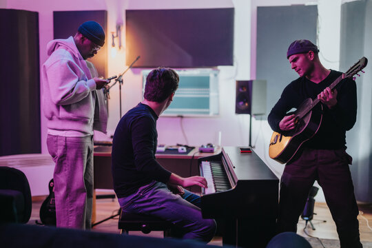 A diverse group of musicians working together in a studio. One plays guitar, another sits at the piano, and the third checks a device. The setting embodies creativity and collaboration.