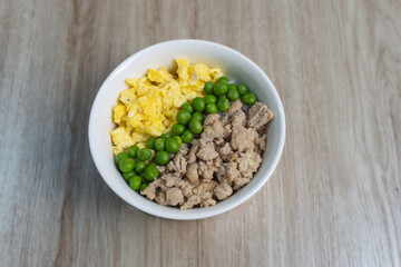Top-down view of a simple and balanced meal in a white bowl, featuring scrambled eggs, green peas, and ground chicken, placed on a wooden table