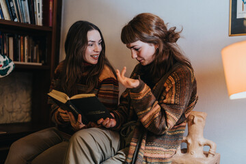 Two young women sitting together indoors, engaging in conversation with a book in hand, evoking a...