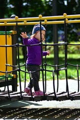 Fototapeta premium Toddler girl balancing on rope bridge at playground, wearing purple jacket and printed hat. Confident expression symbolizes independence, adventure, and motor skill development in outdoor play.