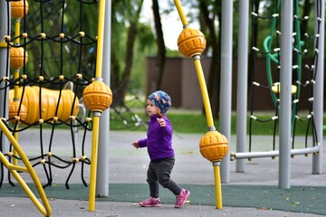 Toddler girl walking confidently on colorful playground, wearing purple jacket and printed hat....