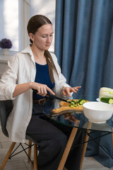 Young woman sitting at a glass table carefully slicing fresh cucumbers on a wooden cutting board,...
