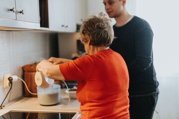 An elderly lady in a red sweater using a mixer in the kitchen while a young man observes. The cozy...