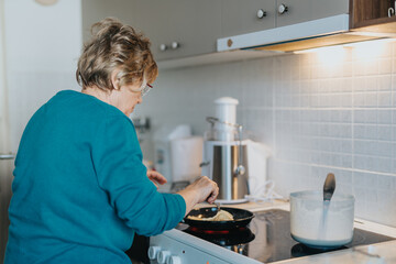 A senior woman cooking in a bright kitchen, mixing ingredients in a frying pan on a stove. A...