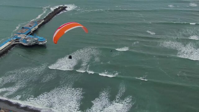 A paraglider flying above the ocean waves near a pier in Miraflores, Lima, Peru