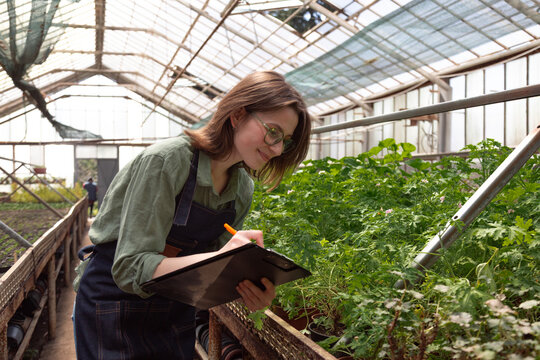 Woman in workwear observing greenery while documenting plant health.