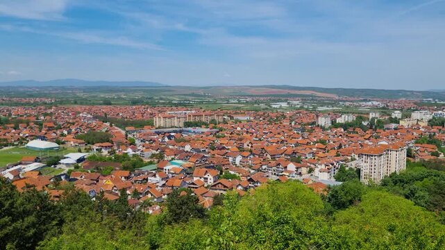 Panoramic view over the red-tiled rooftops of Leskovac in southern Serbia on a sunny spring day, capturing the charm of everyday life and the surrounding hillside beneath a clear blue sky