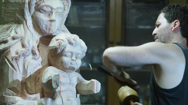 Sculptor powerfully strikes a chisel into a wooden Madonna and Child statue with a mallet, sending chips flying as he carves the unfinished figure still marked by guiding lines and rough surfaces.