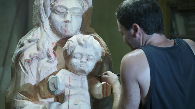 Sculptor carves a large wooden Madonna and Child statue, using a chisel to define forms on the unfinished surface, where red guide lines and rough textures mark the early stage of sacred artistry.