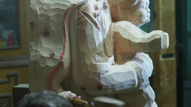 Close-up of artisan using strong mallet blows with a chisel to shape the wood statue of the Virgin and Child, as rough chips fall and major volumes are still marked with red outlines for guidance.