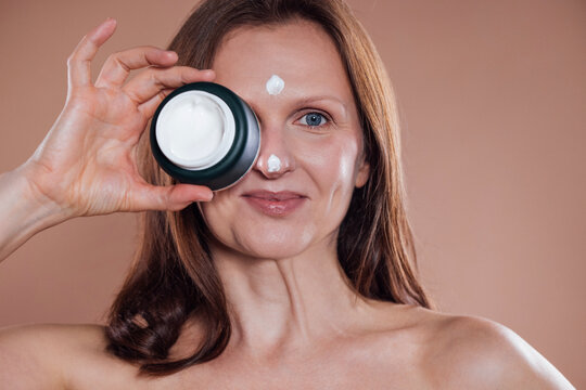 A middle-aged woman holds a jar of cream to her face and smiles. The young lady applied several points of moisturizer to her forehead, nose, and cheek.