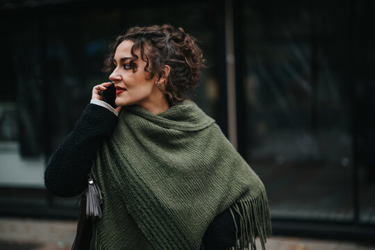 Stylish woman wrapped in a green shawl speaking on her mobile phone in the city. Expressing elegance and modern lifestyle among urban surroundings, the visual embodies casual communication.