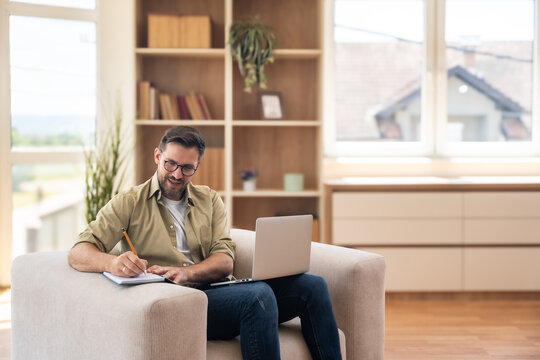Young professional businessman sitting at home in chair working on laptop computers writing ideas in notes. Male business person taking online class, develop independent project working as freelancer. - Powered by Adobe