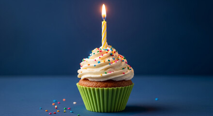 Green cupcake with white frosting and sprinkles, topped with a lit candle against a dark-blue background, symbolizing celebration and joy