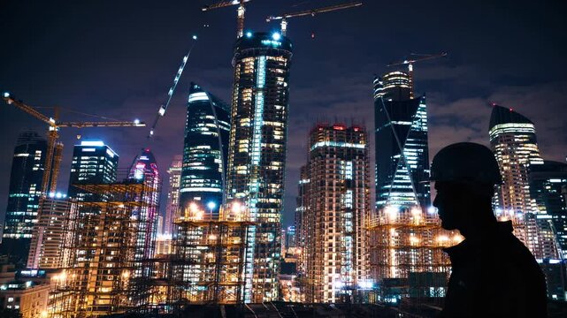 Worker silhouette helmet watches city lighting. Skyscraper towers, cranes and concrete framework shape night architecture. Construction lighting reveals tower, crane, city, concrete and architecture.