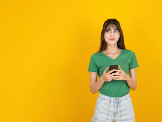 Young woman using phone, portrait of fun smiling positive young woman using phone she wear green t-shirt and jean hold in hand use smartphone look aside copy space promo workspace yellow background.