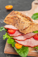 A rustic sandwich made with crusty whole grain bread, layered with slices of smoked pork, fresh spinach leaves, and colorful cherry tomatoes on a wooden board.