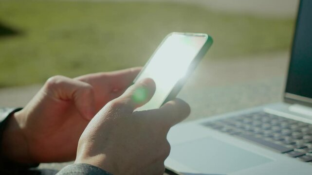 Close-up of female hands using smartphone outdoors with sun glare on screen. Natural light reflections obscure display content. Laptop visible nearby, indicating remote work setting