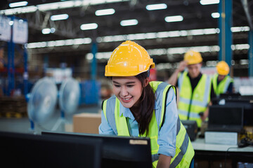 Fototapeta premium Smiling industrial worker in protective helmet using desktop computer in distribution center, focusing on inventory management tasks.