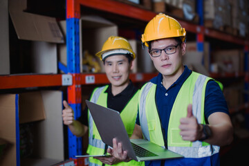 Two Asian warehouse workers in safety helmets and vests give thumbs up while checking data on a laptop, showing teamwork and satisfaction in industrial logistics. Warehouse staff coordination concept