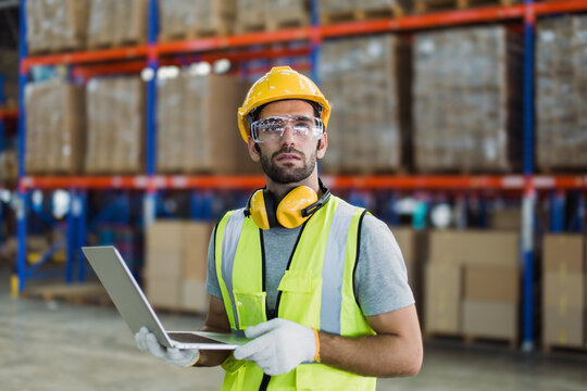 Warehouse logistics coordinator wearing safety gear using laptop to manage transportation schedules and cargo operations in a busy industrial distribution center.