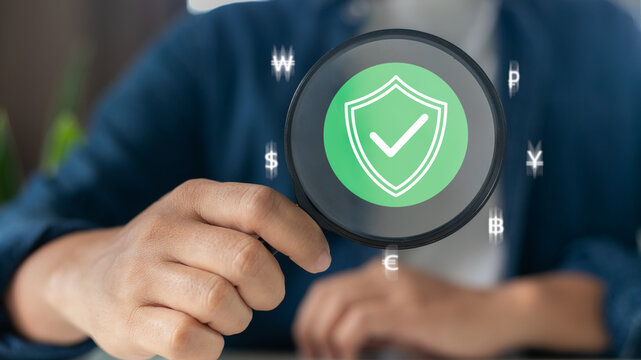 Hand holding magnifying glass with shield and check mark icon, surrounded by currency symbols. Concept of financial security, data protection, secure transaction and fintech.