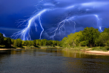 Multiple bright lightning bolts streak across a vivid blue storm sky above a peaceful river surrounded by lush green forest.