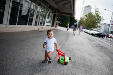 Toddler walking on sidewalk with toy lawnmower and parents in background