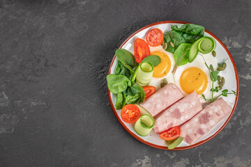 A nutritious breakfast plate with sunny-side-up eggs, deli meat rolls, fresh spinach, cherry tomatoes, cucumber ribbons, and pumpkin seeds sits on a dark textured background
