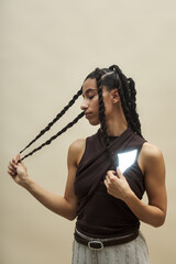 Young adult Black woman standing against plain background holding braided hair with one hand while looking down and to side, wearing sleeveless top and textured skirt