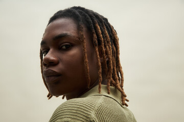 Portrait of young adult Black man turning head toward camera, wearing neutral expression, short dreadlocks visible, standing against plain background, upper body in frame