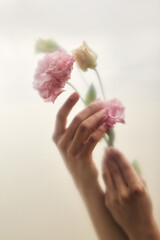 Caucasian young adult woman hands gently holding delicate pink and yellow flowers against soft neutral background, fingers carefully arranging blossoms, focus on graceful gesture