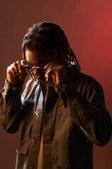 Young adult Black man adjusting sunglasses while standing against plain red background, head...