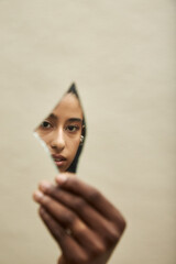 Portrait of young adult Black woman gazing into broken mirror shard, partial face and hand visible,...