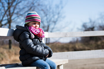 Sad child sitting alone on white bench in winter