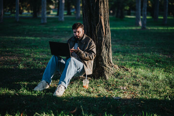 A man enjoys a serene moment, working on his laptop while sitting under a tree in a lush, green...