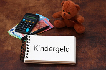 Das Wort Wort Kindergeld auf einen Notizblock geschrieben mit Taschenrechner, Geldscheinen und einem Teddybär.