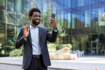 An African American businessman waves while video calling on his smartphone outside a modern office building, smiling and greeting someone on the call.