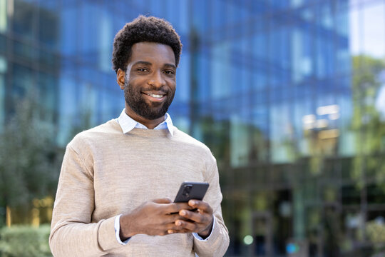 A smiling man stands in front of a modern building, holding a phone.