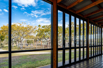Naklejka premium An enclosed patio or covered outdoor seating area with bi-fold glass doors with a view of sunny suburban park with trees and a children's playground outside.