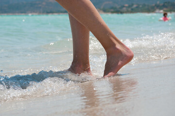 Close-up of female legs walking barefoot along the beach with sea waves. Wellness, travel and lifestyle concept