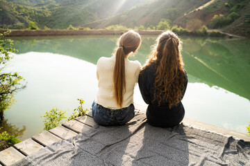Rear view of two young women best friends sitting on wooden pier by mountain lake at sunset hugging looking together at the distance and enjoying peaceful nature, friendship, support, female bonding