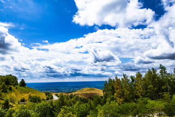 Obraz premium Winding road meanders through lush hills under a bright blue sky filled with fluffy white clouds. A scenic valley opens in the distance, revealing tranquil beauty.