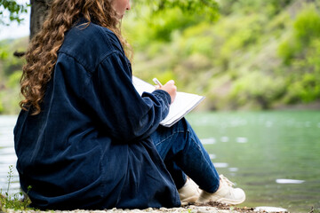 Unrecognizable woman with curly hair writing down thoughts and ideas in personal notebook taking notes journaling keeping manifesting diary as a routine sitting in nature park by lake outdoors