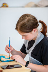 Young artist focused on painting a decorative object with precision. Wearing a protective apron,...