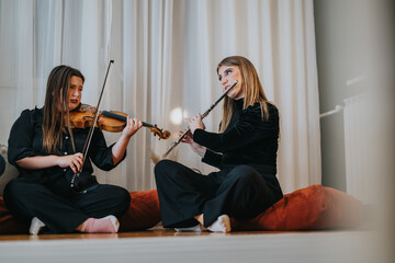Two women musicians are sitting indoors, playing violin and flute together, creating a warm and...
